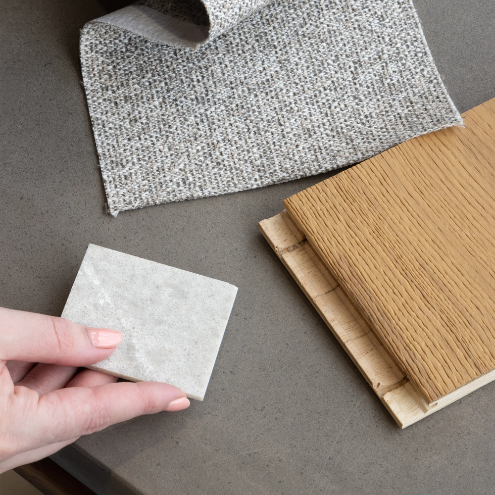 A hand holds a small square sample of light gray textured material on a dark gray countertop, with rolled and stacked material samples nearby, including a wood-look sample.