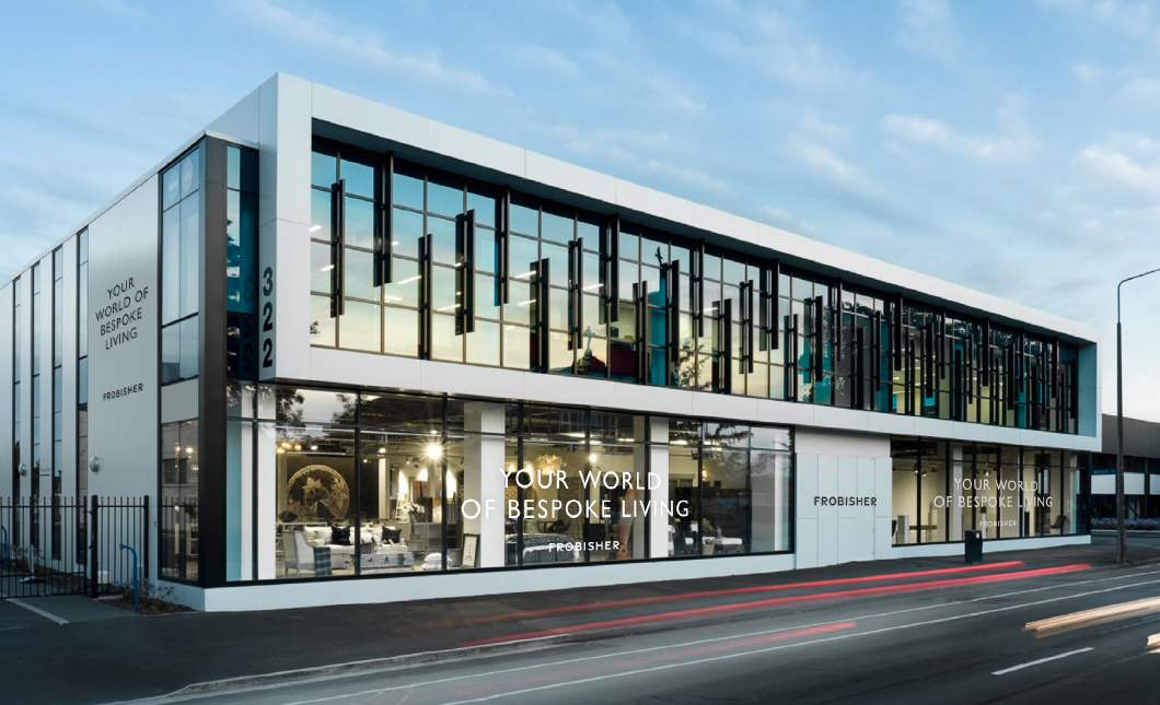 A modern two-story retail showroom building with large glass windows, white exterior walls, and vertical hanging light fixtures, situated on a street with light trails visible on the asphalt under a blue sky.